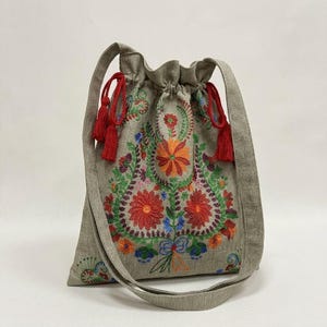 Puede incluir: Un bolso gris con cordón y un diseño de bordado floral colorido. El bordado presenta flores rojas, naranjas, verdes y azules. El bolso tiene una correa para el hombro y borlas rojas.