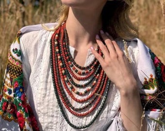 May include: A multi-strand necklace featuring red, black, and tan wooden beads. The necklace is worn with a white embroidered blouse and a colorful floral shawl, creating a traditional folk aesthetic.