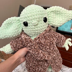 May include: A handmade crochet plush toy of a character with large ears, large black eyes, and a light green complexion. The character is wearing a brown, fuzzy robe. The toy is held in a person's hand.