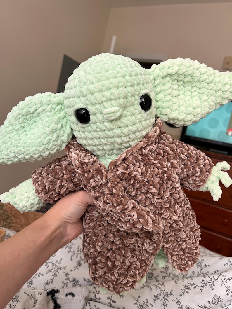 May include: A handmade crochet plush toy of a character with large ears, large black eyes, and a light green complexion. The character is wearing a brown, fuzzy robe. The toy is held in a person's hand.