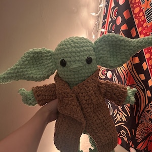 May include: A handmade crochet doll in the likeness of a popular character. The doll features a light green head and large ears, with a brown robe. The doll has black eyes and small hands and feet. The doll is approximately 12 inches tall.