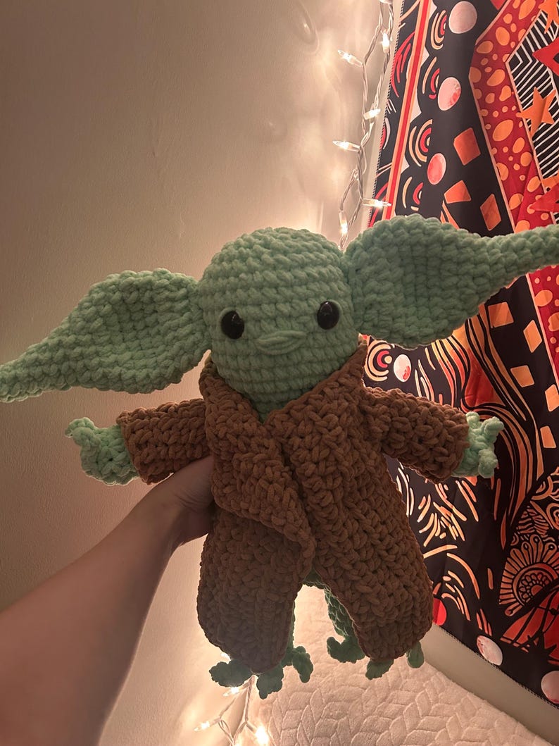 May include: A handmade crochet doll in the likeness of a popular character. The doll features a light green head and large ears, with a brown robe. The doll has black eyes and small hands and feet. The doll is approximately 12 inches tall.