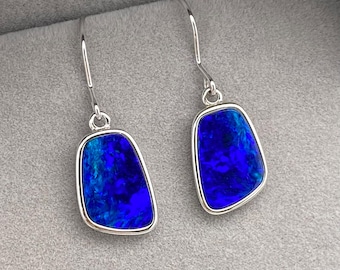 May include: A pair of silver dangle earrings with blue opal stones. The stones are set in a silver bezel and have a slightly irregular shape.