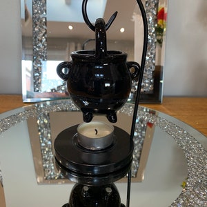Puede incluir: Quemador de aceite de caldero negro con un soporte de metal negro y una vela de té blanca. El quemador está sobre una superficie de espejo con un patrón de diamante.