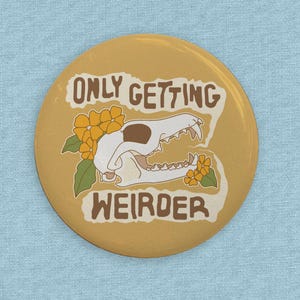 Puede incluir: Un botón amarillo con el texto "ONLY GETTING WEIRDER" en letras marrones. Un cráneo de animal blanco con flores amarillas se encuentra en el centro del botón.