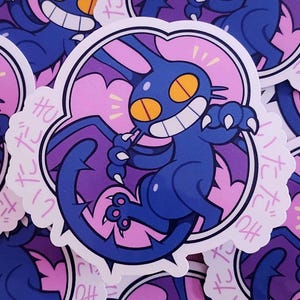 May include: A collection of stickers featuring a cartoonish, dark blue creature with large yellow eyes and a wide grin. The creature has bat-like wings and is set against a pink background with Japanese text. The stickers have a white border.