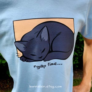 May include: Light blue t-shirt featuring a sleeping black cat illustration inside a peach-colored rectangle. The words "nyap time..." are printed below the cat. The Etsy shop URL is printed at the bottom.