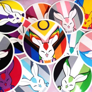 May include: A collection of round stickers featuring a cartoon bunny with various pride flag designs. The stickers showcase a white bunny with a red heart on its head and a happy expression, set against backgrounds of rainbow, transgender, and other pride flag patterns.