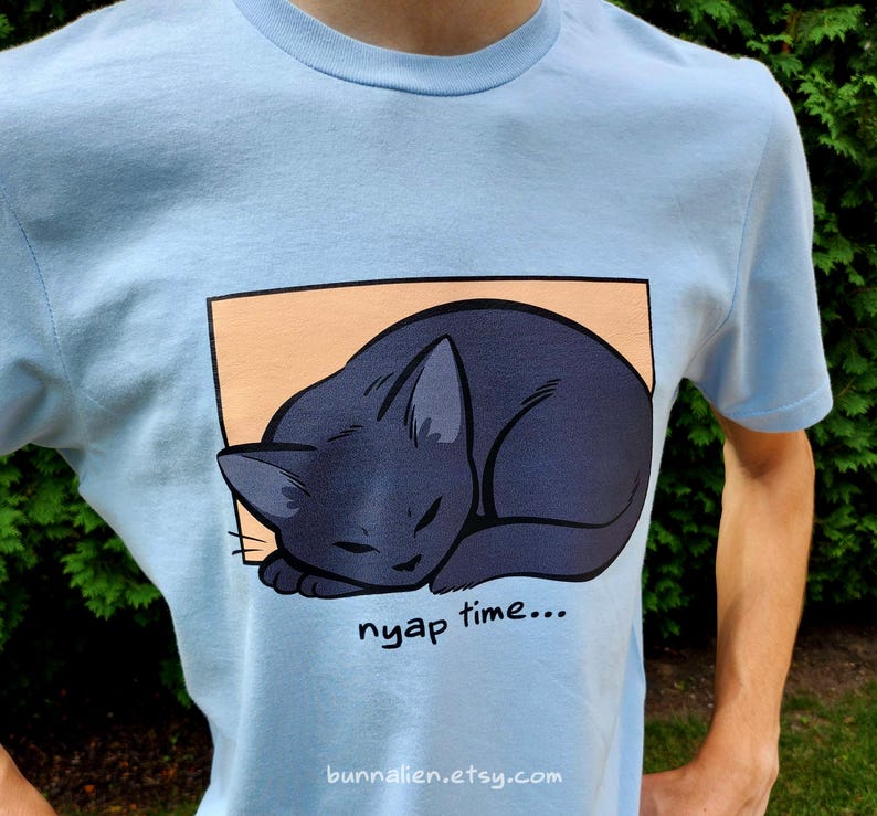 May include: Light blue t-shirt featuring a sleeping black cat illustration inside a peach-colored rectangle. The words "nyap time..." are printed below the cat. The Etsy shop URL is printed at the bottom.