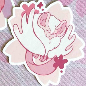 May include: A die-cut sticker featuring a stylized illustration of a white and pink creature, possibly a chinchilla or similar animal, nestled within a pink flower. The design has a soft, pastel color palette.
