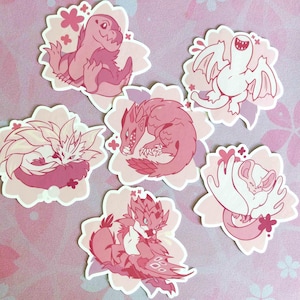 May include: A collection of six pink and white stickers featuring stylized illustrations of fantasy creatures. Each sticker is shaped like a flower with a pink background and a white border. The creatures include dragons and other mythical beings.
