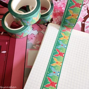 Może przedstawiać: Zielone rolki taśmy washi z powtarzającym się wzorem kolorowych ilustracji królików. Pasek taśmy jest ułożony na notatniku w kratkę. Tło zawiera różowe wzory kwiatowe.