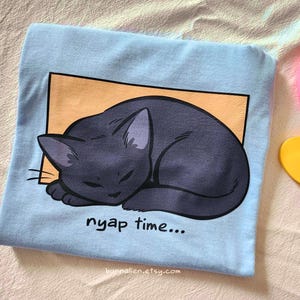 May include: A light blue t-shirt featuring a sleeping black cat illustration within an orange rectangle. The text "nyap time..." is printed below the cat. The Etsy shop URL is visible at the bottom.