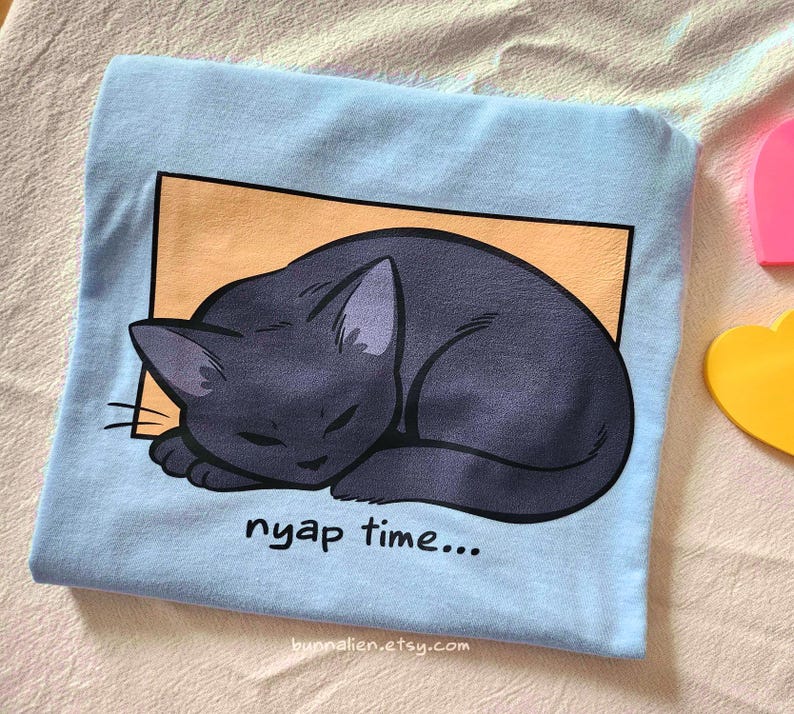 May include: A light blue t-shirt featuring a sleeping black cat illustration within an orange rectangle. The text "nyap time..." is printed below the cat. The Etsy shop URL is visible at the bottom.