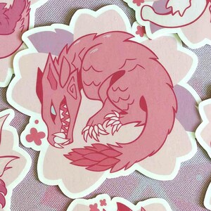 May include: Pink sticker featuring a stylized dragon illustration. The dragon is curled up, with sharp claws, teeth, and a spiky mane. The sticker is on a light pink floral background, with other similar stickers visible.