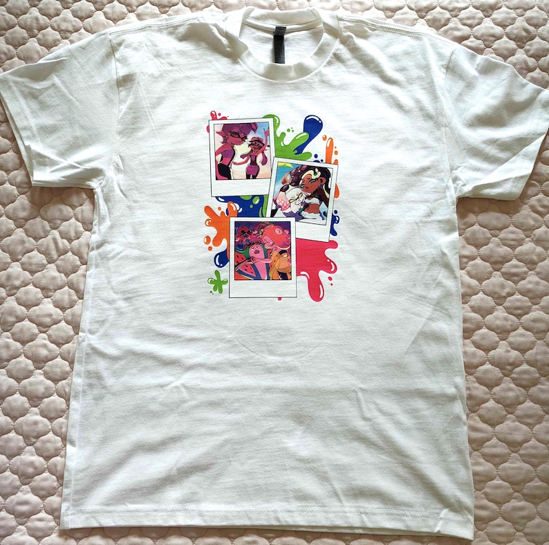 Puede incluir: Una camiseta blanca de manga corta con un estampado gr&aacute;fico de tres fotos tipo polaroid apiladas, rodeadas de salpicaduras de pintura de colores en tonos azul, verde, naranja y rosa. Las fotos muestran personajes animados en varias escenas.