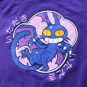 Può includere: T-shirt viola con un'illustrazione a cartoni animati di una creatura blu con occhi gialli e artigli affilati. Il design include una forma di nuvola rosa e viola e testo giapponese. La creatura ha una coda appuntita.