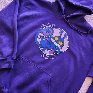 May include: A purple hoodie with a front pocket and drawstring hood. The front features a cartoon illustration of a blue creature with yellow eyes and the Japanese text "Itadakimasu" in white.