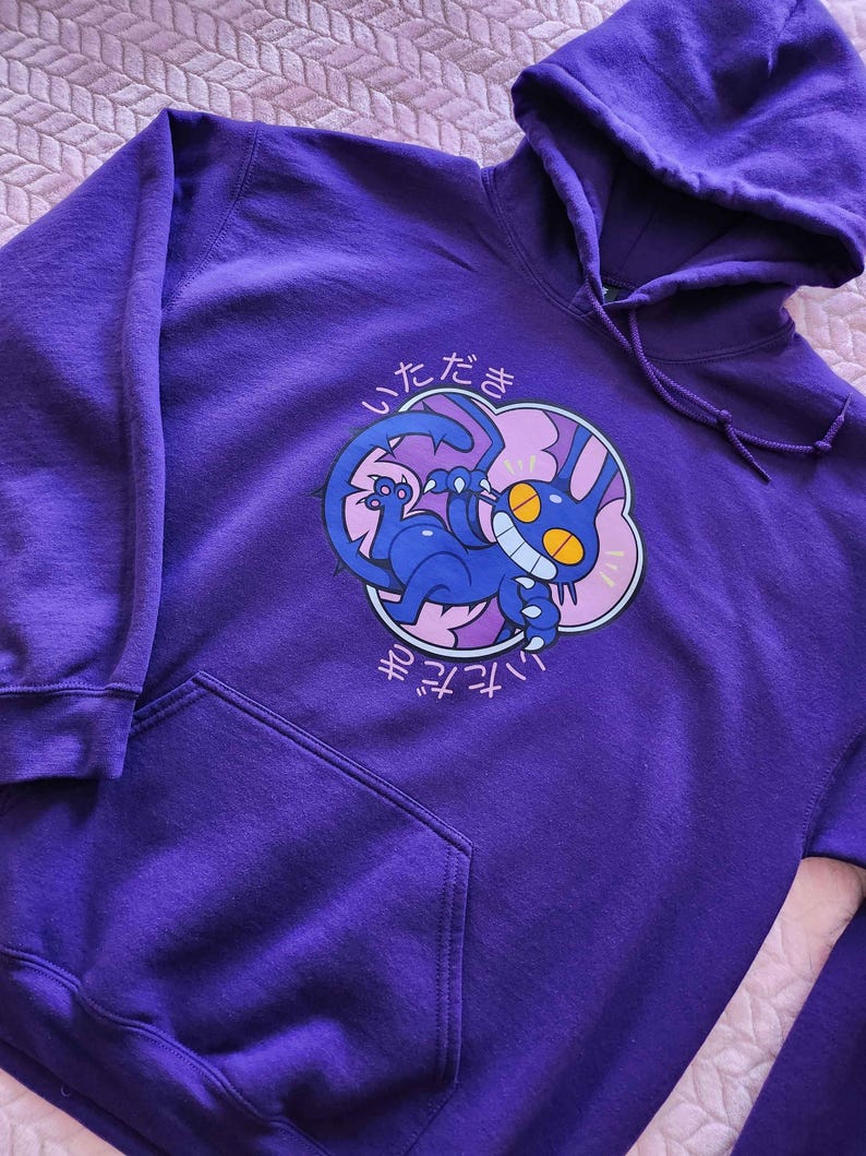 May include: A purple hoodie with a front pocket and drawstring hood. The front features a cartoon illustration of a blue creature with yellow eyes and the Japanese text "Itadakimasu" in white.