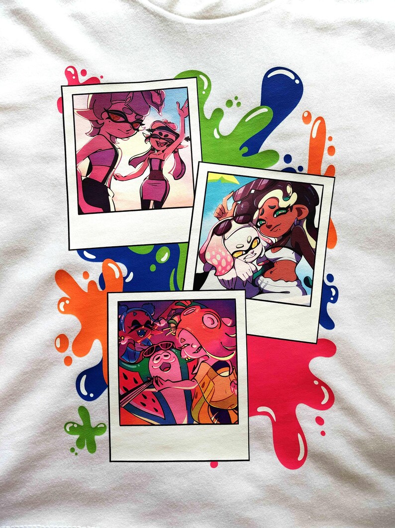 Puede incluir: Una camiseta blanca con un estampado gr&aacute;fico de tres fotos superpuestas al estilo polaroid, rodeadas de salpicaduras de pintura de colores. Las fotos muestran personajes animados en diversos entornos, incluyendo escenas de playa.