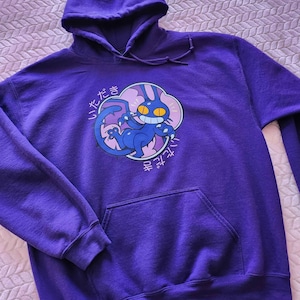 May include: A purple hooded sweatshirt with a front pocket and drawstring hood. The sweatshirt features a cartoon graphic of a blue cat-like creature with yellow eyes and Japanese text. The graphic is set within a pink cloud shape.