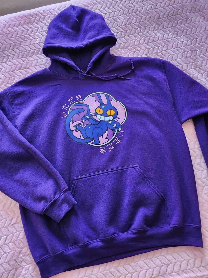 May include: A purple hooded sweatshirt with a front pocket and drawstring hood. The sweatshirt features a cartoon graphic of a blue cat-like creature with yellow eyes and Japanese text. The graphic is set within a pink cloud shape.