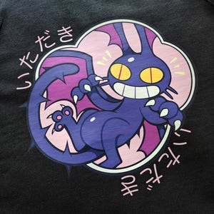 May include: Black tank top featuring a cartoon creature with a purple body, yellow eyes, and a wide grin. The creature is surrounded by a pink and purple cloud-like shape with Japanese text. The design is centered on the front of the tank top.
