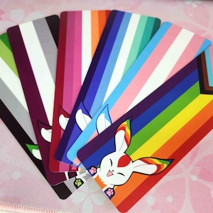 May include: A set of ten colorful bookmarks featuring a rainbow design with a white rabbit illustration. The bookmarks are made of cardstock and have a glossy finish.