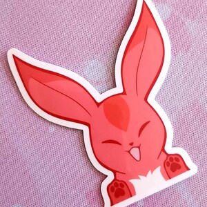 May include: A red cartoon bunny sticker with a white border. The bunny has large ears, a smiling face, and paw prints on its paws. The sticker is on a pink background.