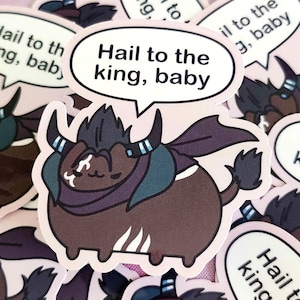 Peut inclure: Autocollant représentant un taureau de dessin animé avec un corps marron foncé, une cape violette et des cornes. Le taureau a une bulle de dialogue avec le texte "Hail to the king, baby" en noir. Fond rose.
