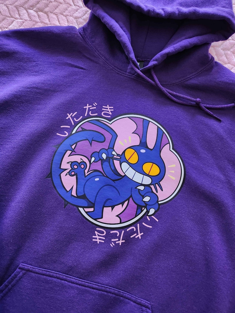 May include: Purple hooded sweatshirt featuring a cartoon cat-like creature with yellow eyes and a toothy grin. The graphic is surrounded by a pink cloud and Japanese text. The hoodie has a front pocket and drawstring.