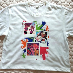 Puede incluir: Una camiseta blanca de manga corta con un estampado gr&aacute;fico de tres fotos superpuestas al estilo polaroid. Las im&aacute;genes muestran personajes de dibujos animados coloridos, rodeados de salpicaduras de pintura azul, verde y naranja.
