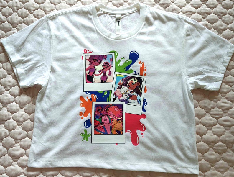 Puede incluir: Una camiseta blanca de manga corta con un estampado gr&aacute;fico de tres fotos superpuestas al estilo polaroid. Las im&aacute;genes muestran personajes de dibujos animados coloridos, rodeados de salpicaduras de pintura azul, verde y naranja.