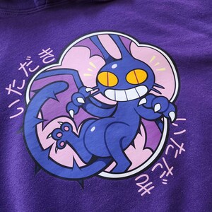 May include: Purple hoodie featuring a cartoon creature with a large tail, claws, and yellow eyes. The creature is dark blue with a white-toothed grin, set against a pink and white cloud-like shape. Japanese text is printed around the design.