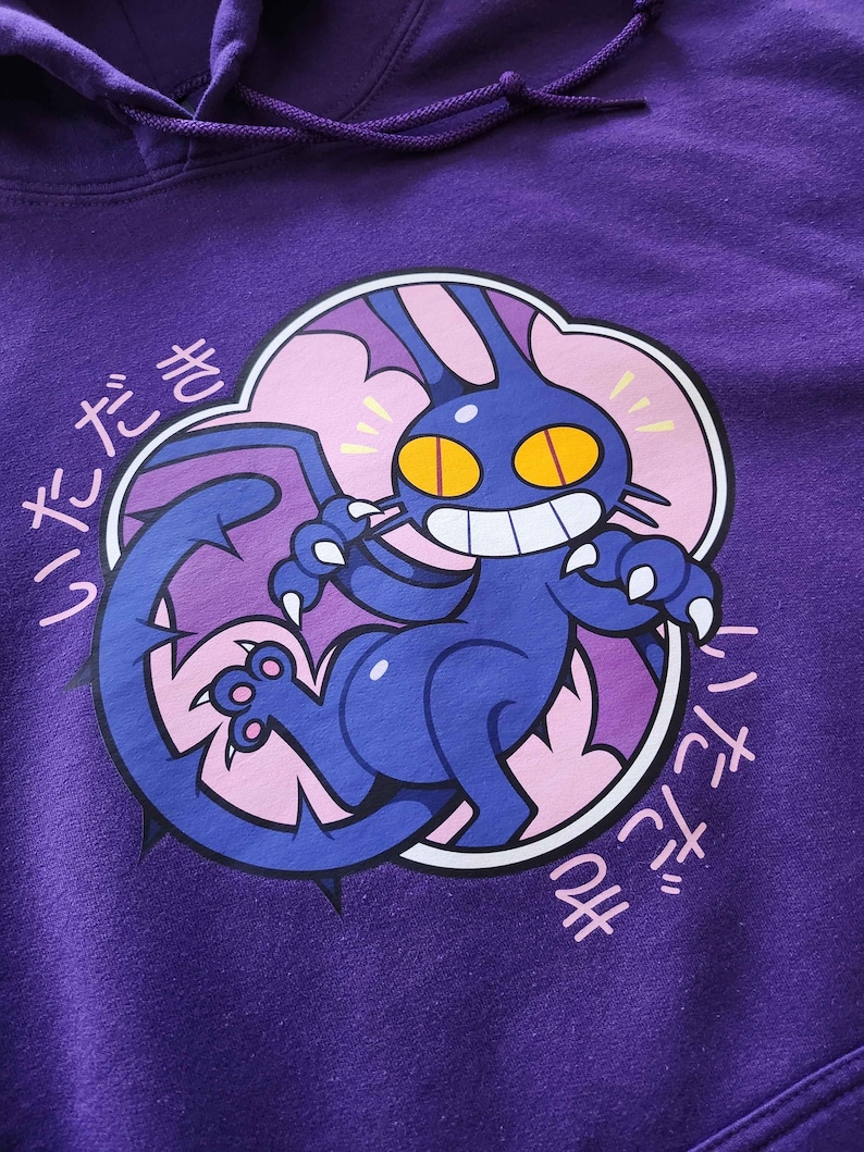 May include: Purple hoodie featuring a cartoon creature with a large tail, claws, and yellow eyes. The creature is dark blue with a white-toothed grin, set against a pink and white cloud-like shape. Japanese text is printed around the design.