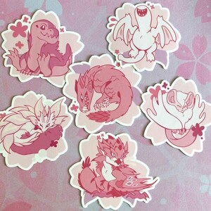 May include: A collection of six pink and white stickers featuring cartoon monster illustrations. Each sticker is shaped like a flower and has a different monster design, including dragons and other fantasy creatures. The stickers are in various shades of pink.