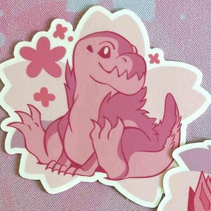 May include: A pink dinosaur sticker with a flower-shaped border. The dinosaur is sitting with its arms out and has a friendly expression. The sticker has a light pink and dark pink color scheme.