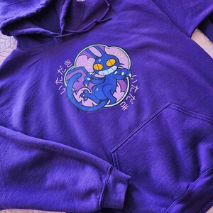 May include: A purple hoodie with a drawstring hood and a kangaroo pocket. The front features a graphic of a blue cat with yellow eyes and a mischievous grin, surrounded by Japanese text. The hoodie is made of a soft, comfortable material.