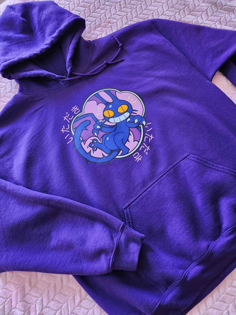 May include: A purple hoodie with a drawstring hood and a kangaroo pocket. The front features a graphic of a blue cat with yellow eyes and a mischievous grin, surrounded by Japanese text. The hoodie is made of a soft, comfortable material.
