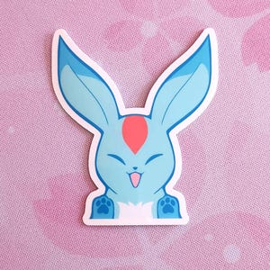 May include: A die-cut sticker featuring a cartoon bunny with large blue ears and a light blue body. The bunny has a red marking on its forehead, a smiling face, and paw prints on its feet. The sticker has a white border.