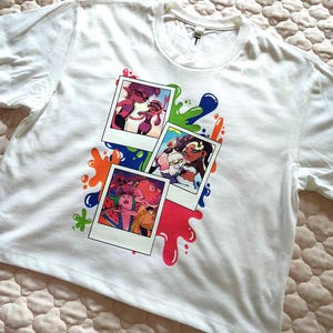 Puede incluir: Una camiseta blanca de manga corta con un estampado gr&aacute;fico de tres fotos superpuestas al estilo polaroid. Las im&aacute;genes muestran personajes animados en entornos coloridos, rodeados de vibrantes salpicaduras de pintura azul, verde, naranja y rosa.