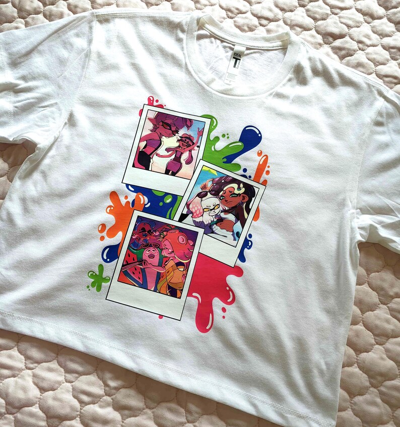 Puede incluir: Una camiseta blanca de manga corta con un estampado gr&aacute;fico de tres fotos superpuestas al estilo polaroid. Las im&aacute;genes muestran personajes animados en entornos coloridos, rodeados de vibrantes salpicaduras de pintura azul, verde, naranja y rosa.