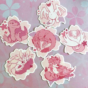 May include: A collection of six pink and white stickers featuring stylized illustrations of fantasy creatures. Each sticker is shaped like a flower and has a different creature design, including dragons and other mythical beings.