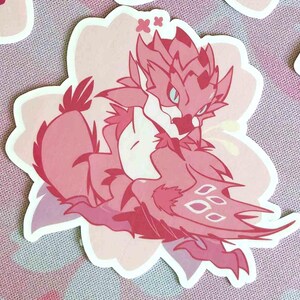 May include: A pink sticker featuring a stylized dragon-like creature with a flower-shaped border. The creature has a detailed design with various shades of pink and white, with a few small stars.