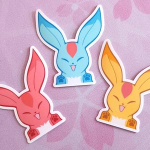 May include: Three colorful bunny stickers. One is red, one is blue, and one is yellow. Each sticker has a white outline and features a smiling bunny face with long ears and paw prints.