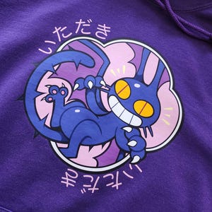 May include: Purple hoodie featuring a cartoon cat design. The cat is blue with yellow eyes and a wide grin, surrounded by a pink cloud-like shape. Japanese text is above and below the graphic. The hoodie has a drawstring.