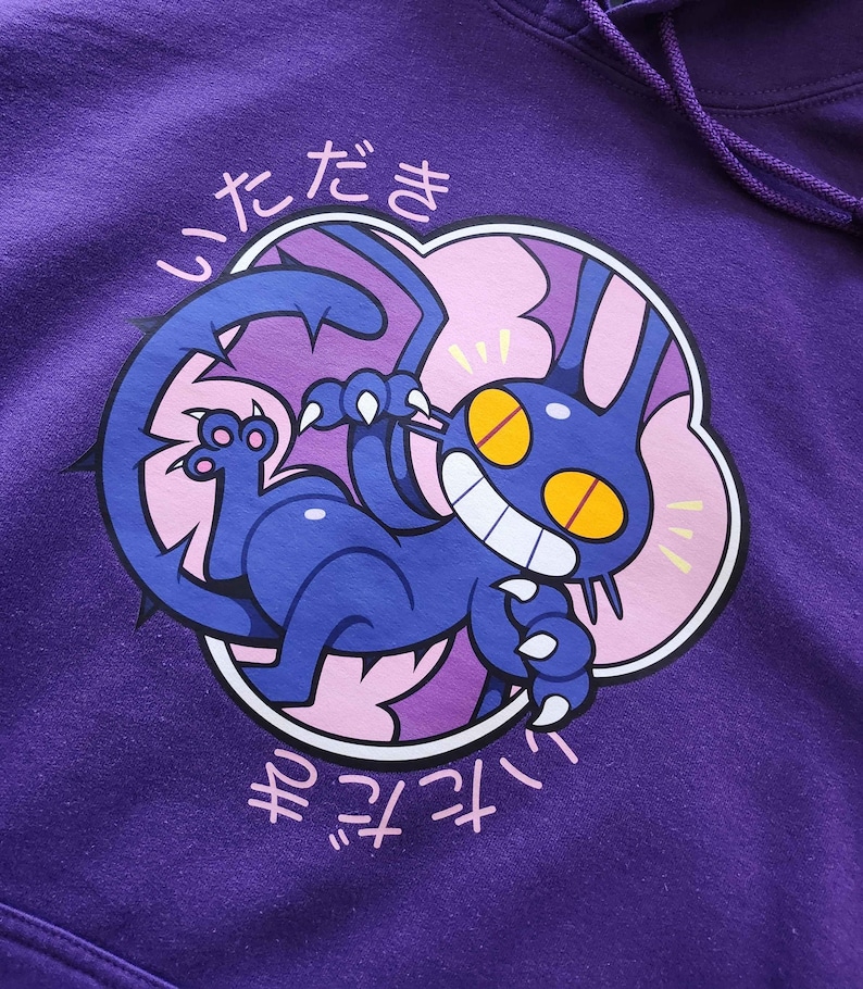 May include: Purple hoodie featuring a cartoon cat design. The cat is blue with yellow eyes and a wide grin, surrounded by a pink cloud-like shape. Japanese text is above and below the graphic. The hoodie has a drawstring.