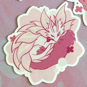 May include: A sticker featuring a stylized, curled-up fox-like creature with a floral mane and tail. The design is primarily pink and white, with a light pink floral-shaped border and small flower accents.