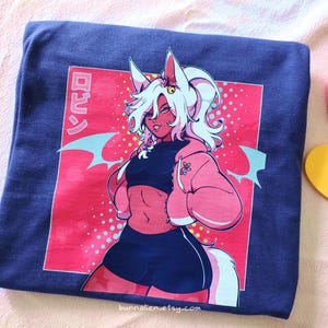 Pu&ograve; includere: Una t-shirt blu navy piegata con un'illustrazione colorata in stile anime. La grafica raffigura un personaggio con capelli bianchi, dettagli rosa e una giacca rosa, su sfondo rosso con testo giapponese. Il nome del negozio Etsy &egrave; visibile.