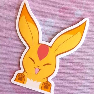May include: A die-cut sticker featuring a cartoon bunny with yellow fur, long ears, and a red heart-shaped marking on its forehead. The bunny has a happy expression with its tongue sticking out and paw prints on its paws.